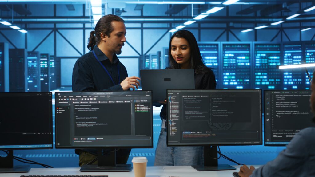 Two professionals review code on a laptop in a tech workspace, surrounded by multiple computer screens displaying programming details in a data center.