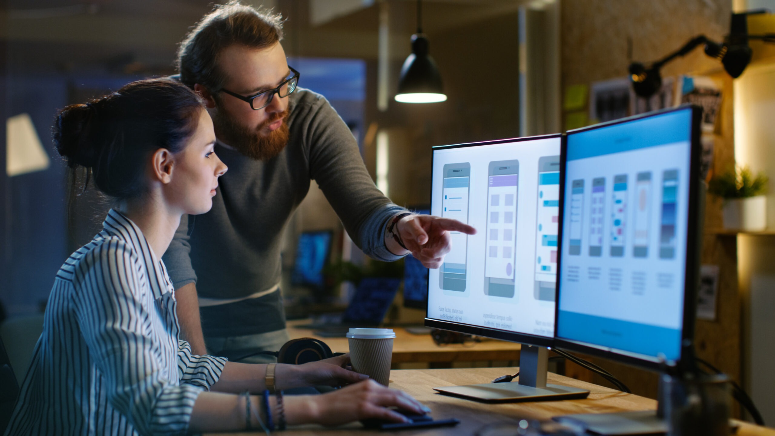 A man points at smartphone app designs displayed on a monitor while a woman watches intently, collaborating in a tech workspace.