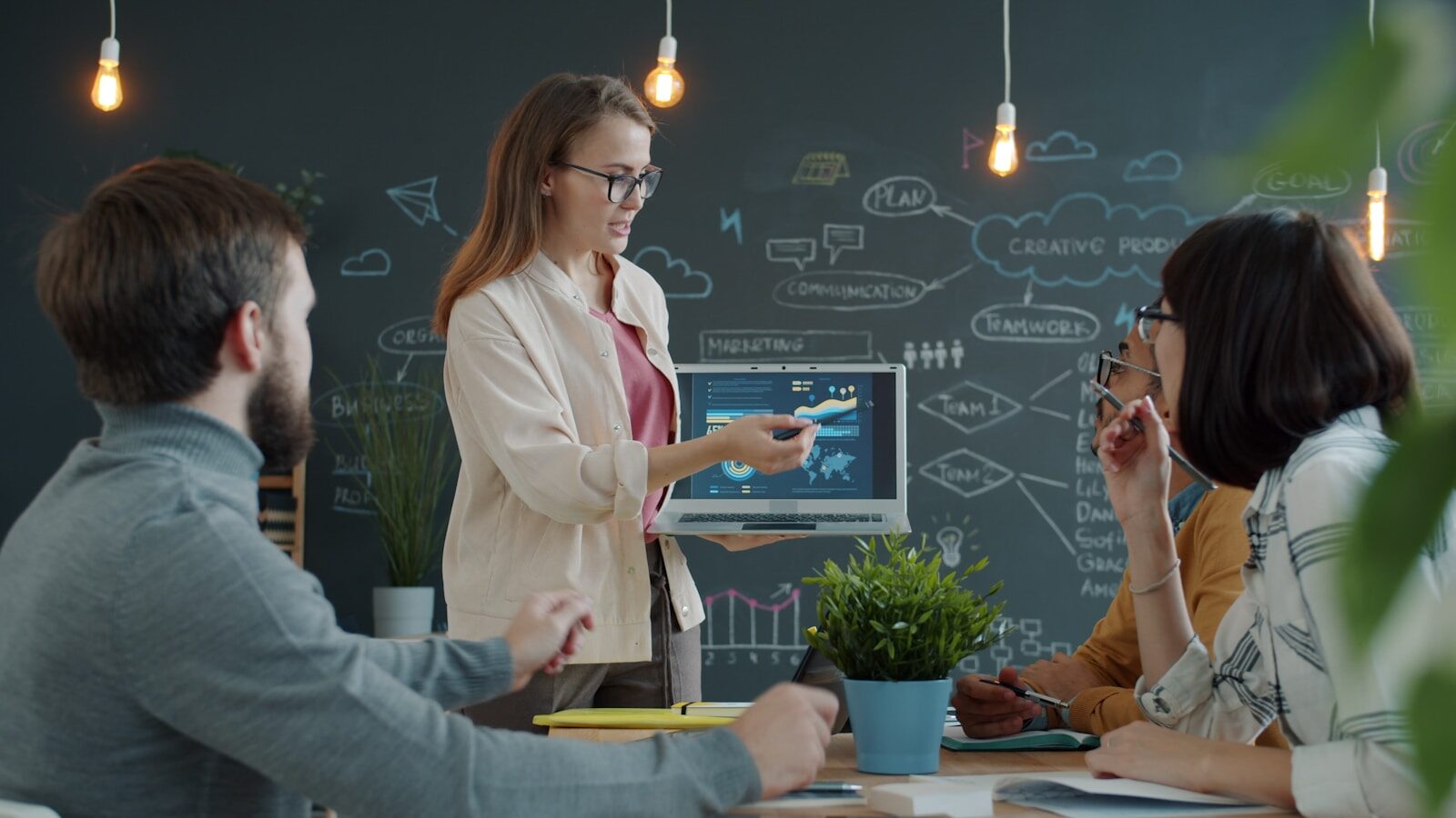 A woman presents data on a laptop to colleagues during a business meeting, with a chalkboard filled with brainstorming ideas in the background.