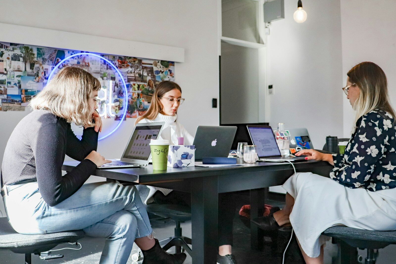 Three women collaborate in a modern office setting, focused on their laptops surrounded by a vibrant wall collage and creative decor.