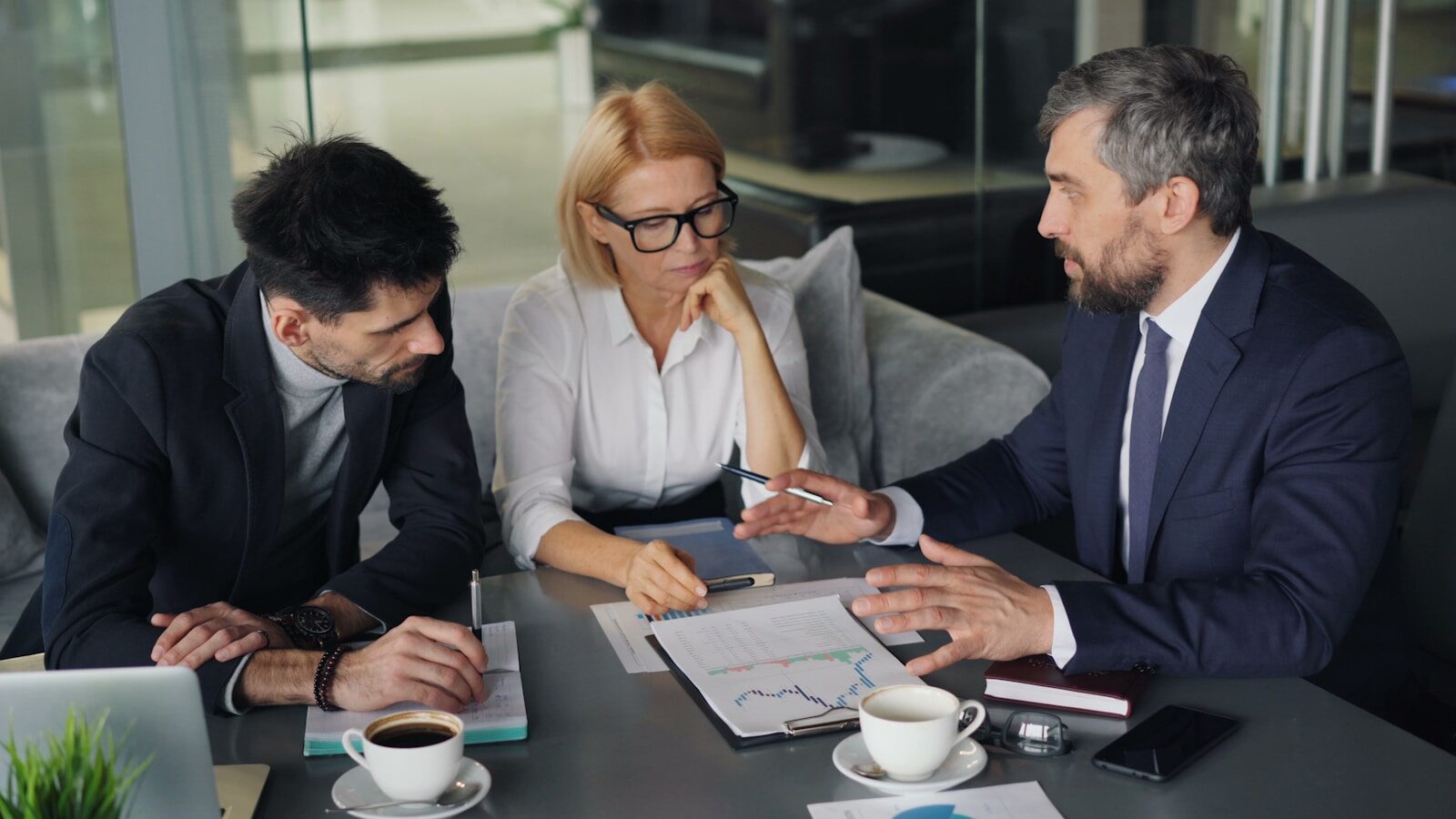 Three professionals engage in a focused discussion around a table with documents and drinks, highlighting a collaborative meeting atmosphere.