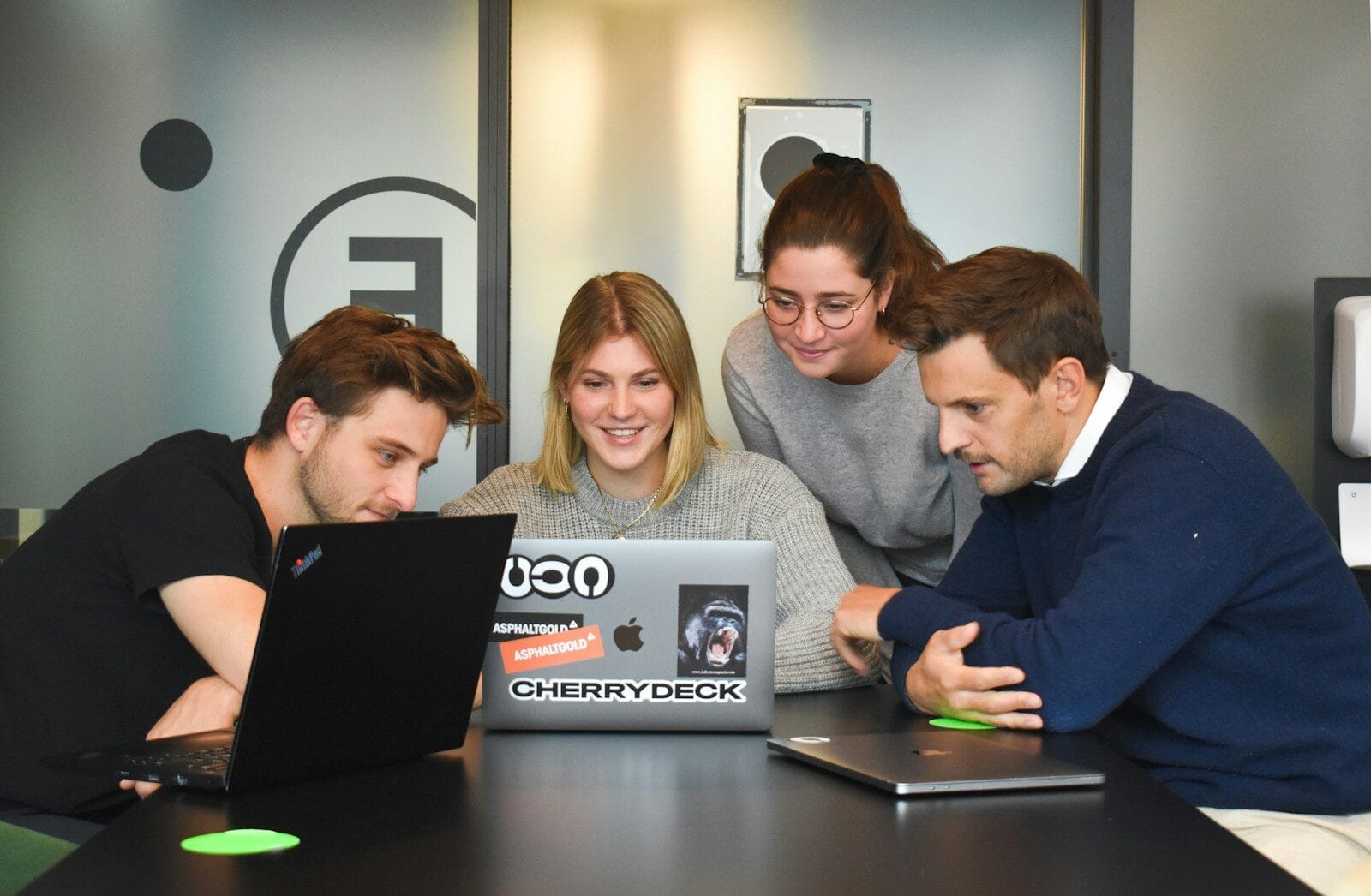 Four coworkers collaborate at a table, focused on a laptop with stickers, sharing ideas and engaging in a creative discussion.